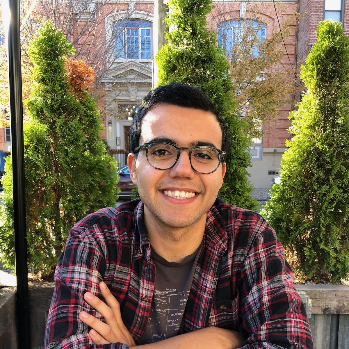 An image of Danny Ayoub, a man with short black hair and glasses, smiling at the camera sitting at a brown wooden table. His elbows are resting on the table and his arms are crossed. A few tall landscaping trees stand in the background.